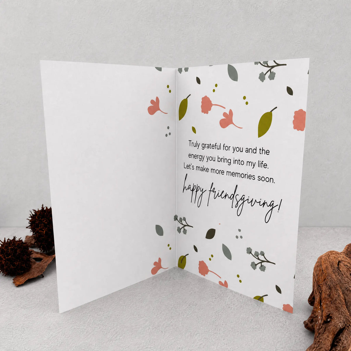 An open white greeting card with a floral pattern and the handwritten message 'happy friendsgiving!' displayed on the right panel, styled on a light gray surface with decorative pinecones and wood in the corners.