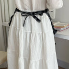 White dress with a black belt worn by a person, with a blurred indoor background.