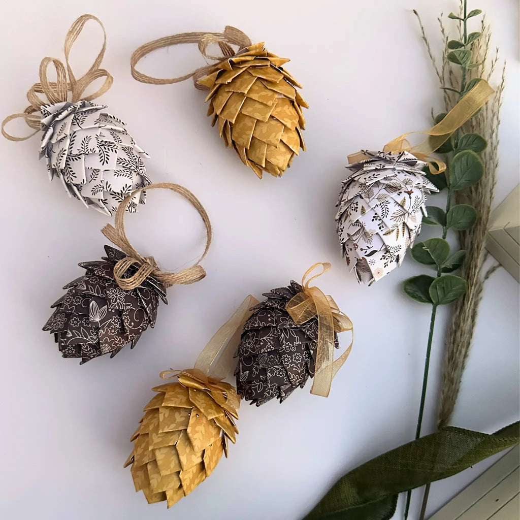 Six decorative pinecone ornaments made from patterned paper in gold, silver, and dark brown tones, arranged on a white background with dried grass and green leaves.