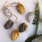 Six decorative pinecone ornaments made from patterned paper in gold, silver, and dark brown tones, arranged on a white background with dried grass and green leaves.
