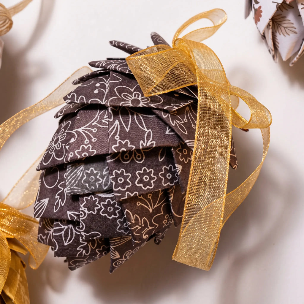 A close-up of a decorative pinecone-shaped ornament made from layered brown fabric petals with a white floral pattern, tied with a sheer gold ribbon on a light background.