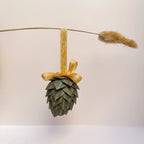 A pinecone-shaped hanging ornament made from layered green paper with gold glitter details, tied to a gold ribbon and displayed on a thin wire with a dried grass stem against a plain light background.