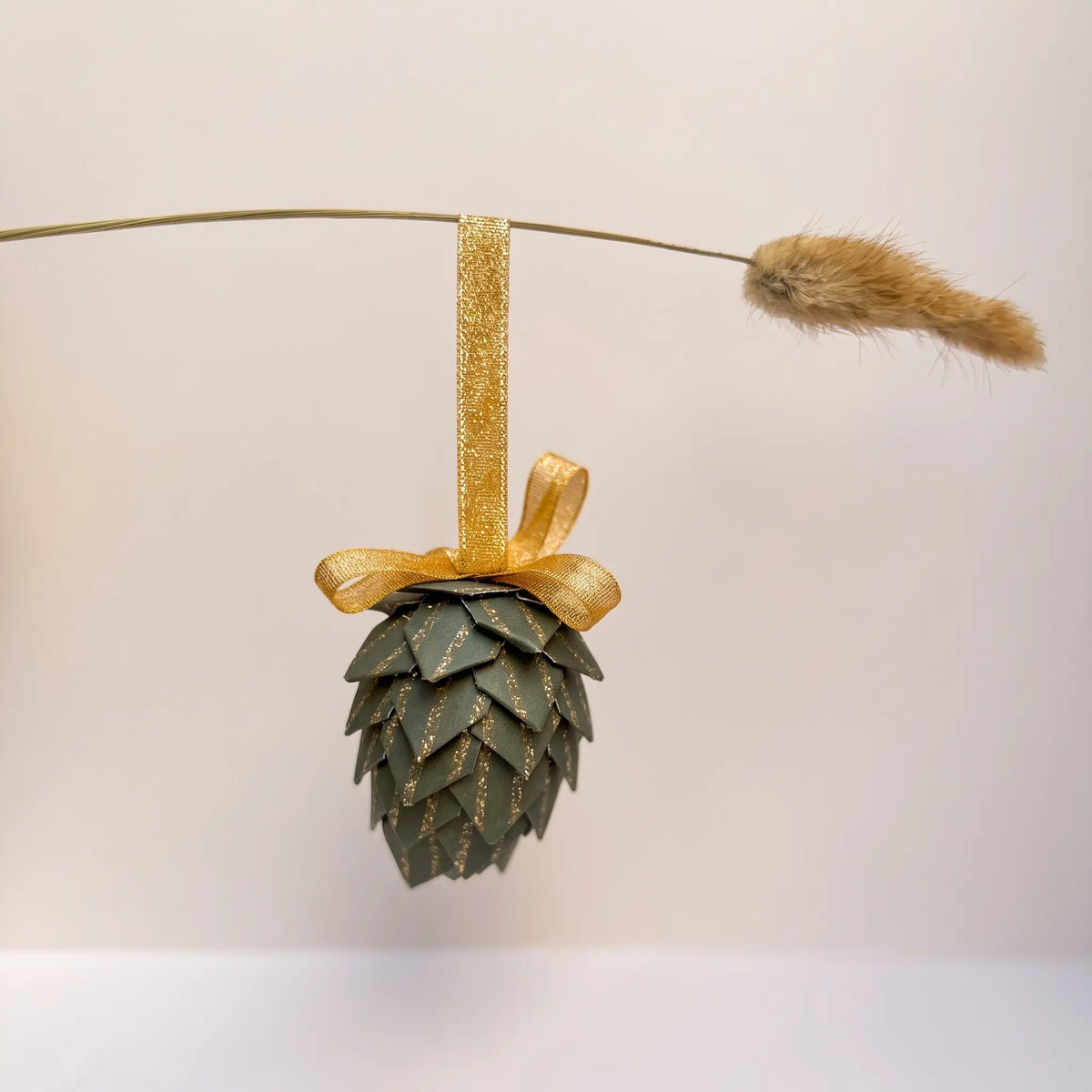 A pinecone-shaped hanging ornament made from layered green paper with gold glitter details, tied to a gold ribbon and displayed on a thin wire with a dried grass stem against a plain light background.