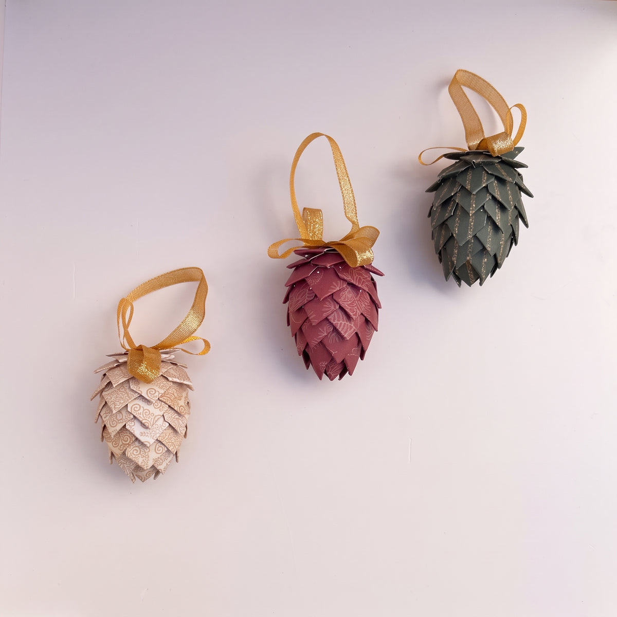 Three pinecone-shaped hanging ornaments in beige, dusty rose, and dark green, each with a gold ribbon loop, displayed on a light off-white background.