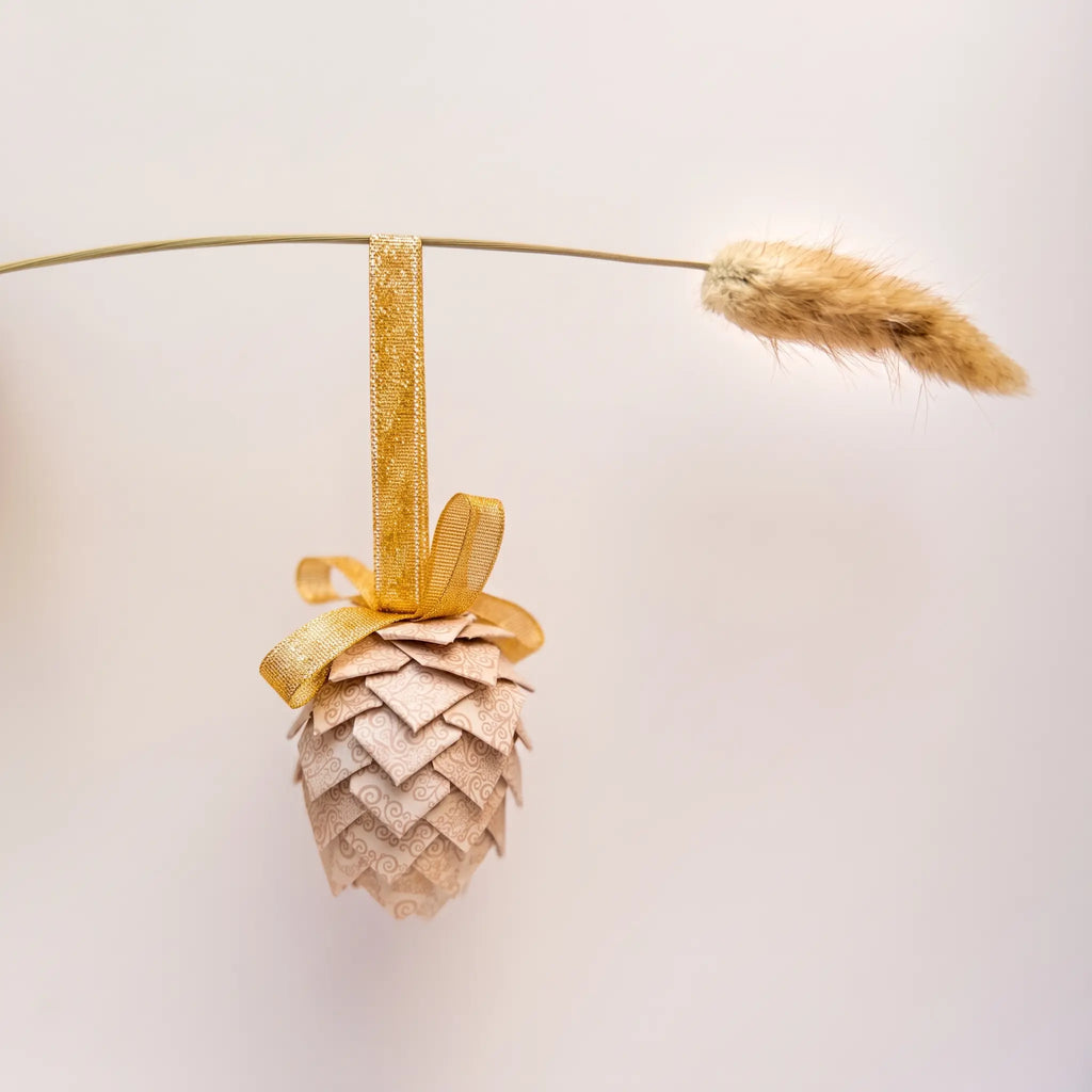 A decorative pinecone-shaped ornament in a light beige tone with a gold ribbon loop and bow, hanging from a thin wire against a plain light background. 