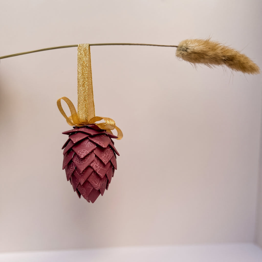 A decorative hanging ornament shaped like a pine cone, made from layered burgundy leather scales, suspended from a gold ribbon and bow. A dried grass stem hangs horizontally in the background against a plain light wall.