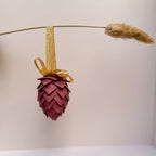 A decorative hanging ornament shaped like a pine cone, made from layered burgundy leather scales, suspended from a gold ribbon and bow. A dried grass stem hangs horizontally in the background against a plain light wall.