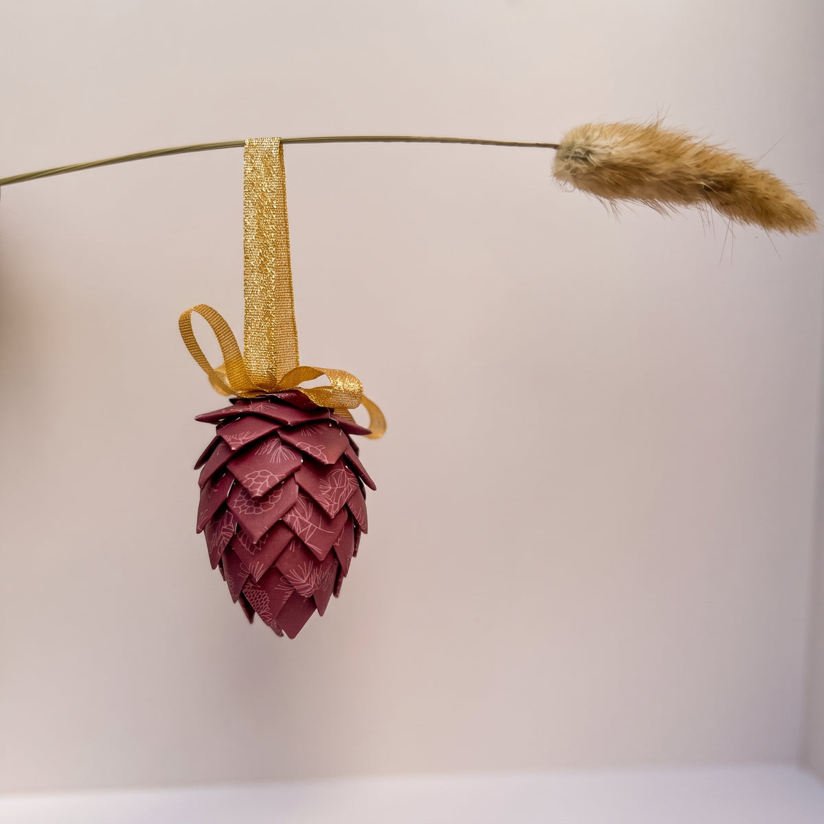 A decorative hanging ornament shaped like a pine cone, made from layered burgundy leather scales, suspended from a gold ribbon and bow. A dried grass stem hangs horizontally in the background against a plain light wall.