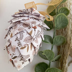 A handmade paper pinecone ornament with a botanical leaf pattern in brown, beige, and muted pink on a white background, decorated with a gold mesh ribbon. It is styled next to a green artificial plant stem.