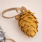A decorative pinecone-shaped ornament made from overlapping gold-toned paper scales, tied with a natural jute loop, photographed on a plain light beige background.