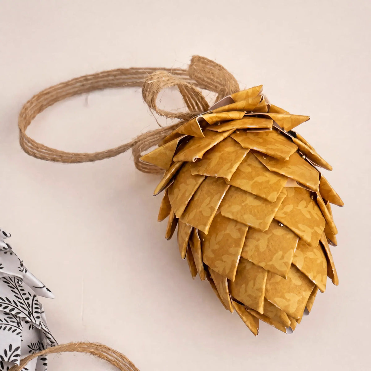 A decorative pinecone-shaped ornament made from overlapping gold-toned paper scales, tied with a natural jute loop, photographed on a plain light beige background.
