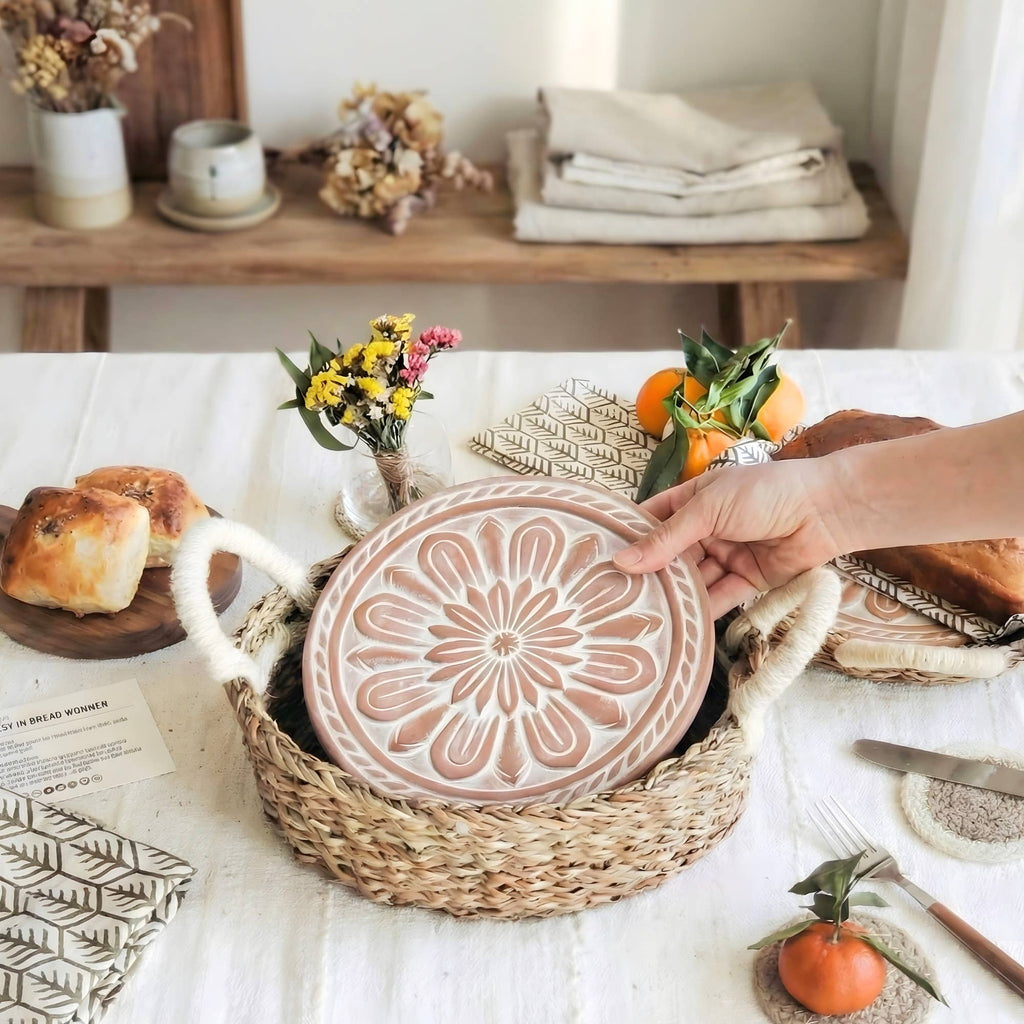 Person holding a decorative plate with a floral design on a table with bread and flowers.