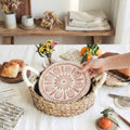 Person holding a decorative plate with a floral design on a table with bread and flowers.