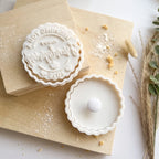 A white ceramic mooncake cutter and matching tray set styled on light wood blocks with scattered powdered sugar and dried grass accents.