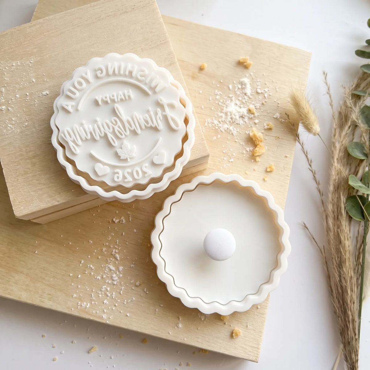 A white ceramic mooncake cutter and matching tray set styled on light wood blocks with scattered powdered sugar and dried grass accents.