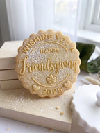 A scalloped round sugar cookie with embossed text reading 'Wishing You Happy Friendsgiving 2026' and a maple leaf design, dusted with powdered sugar and displayed on wooden blocks.