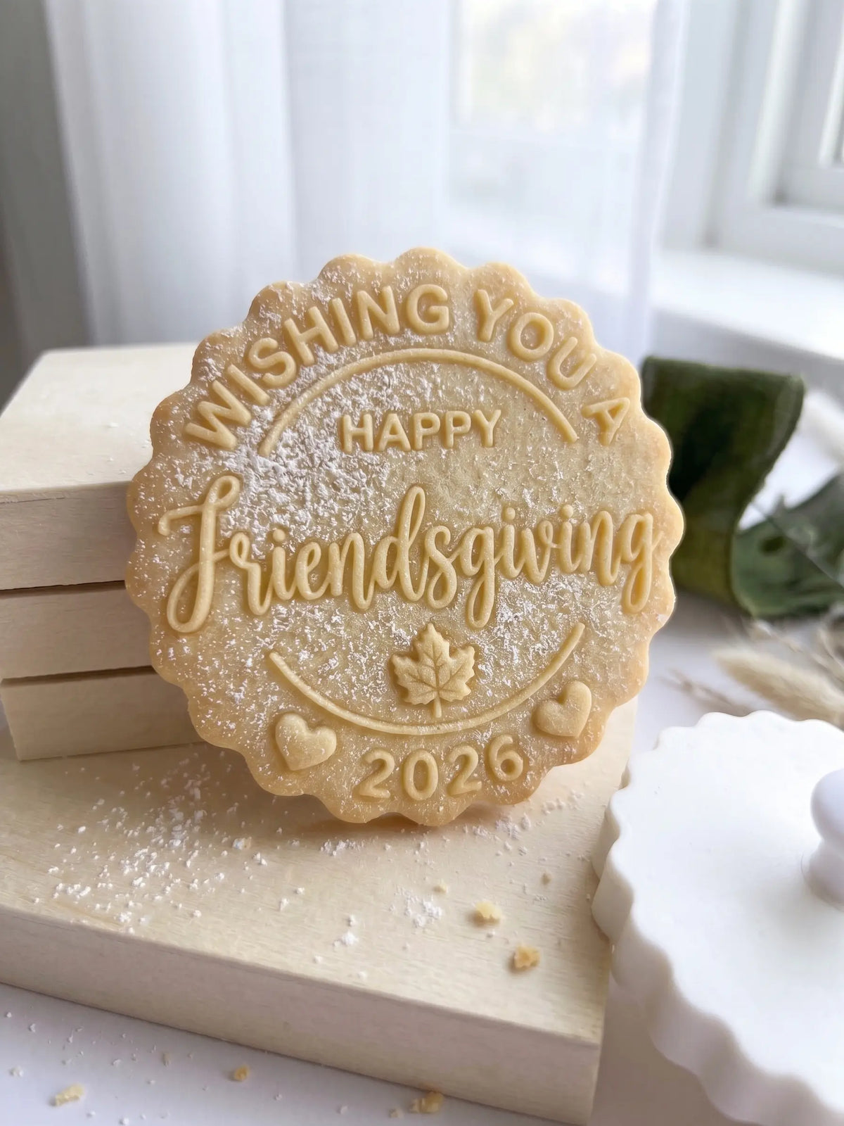A scalloped round sugar cookie with embossed text reading 'Wishing You Happy Friendsgiving 2026' and a maple leaf design, dusted with powdered sugar and displayed on wooden blocks.