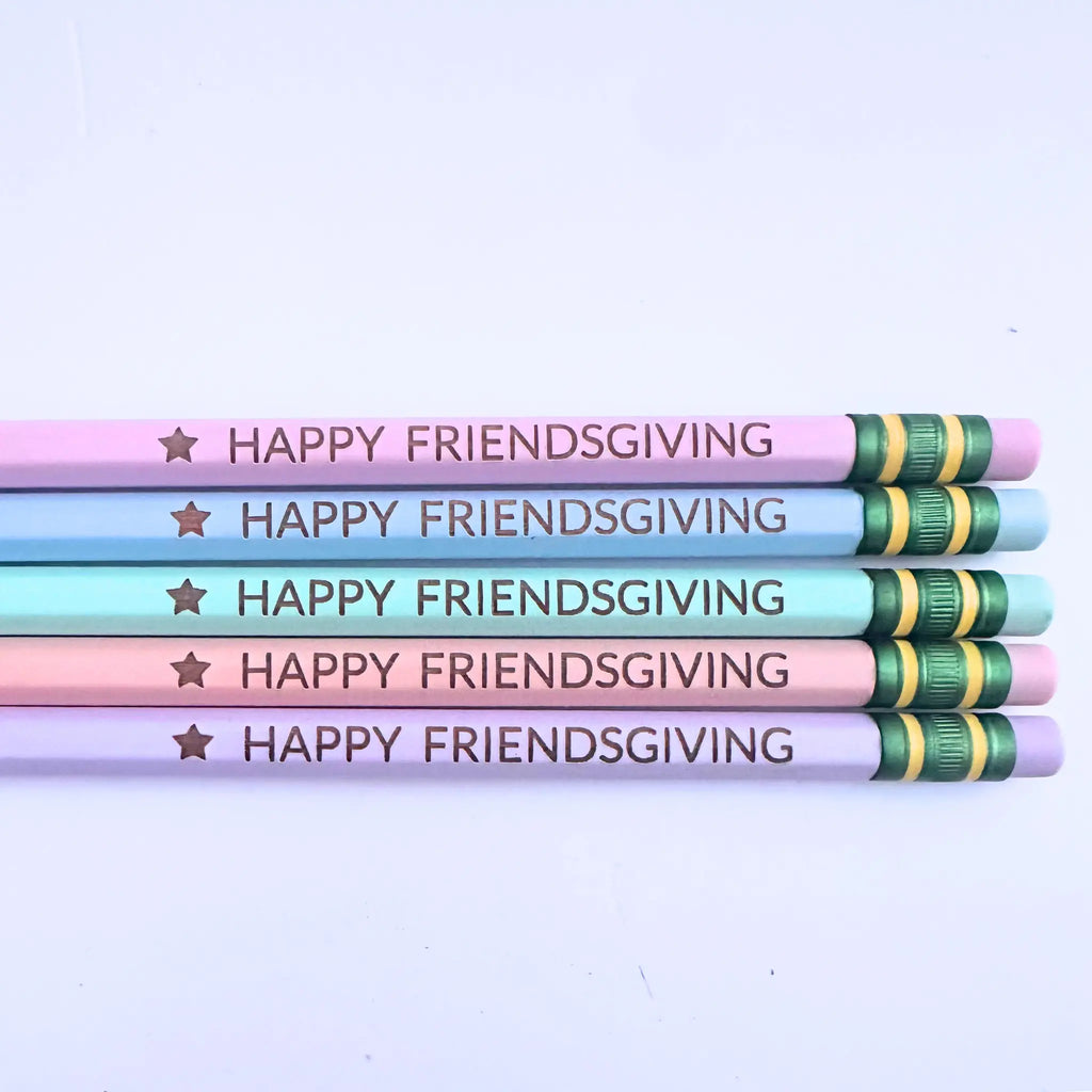 Five decorative pencils stacked horizontally on a light background. The pencils are pastel pink, light blue, and lavender, each printed with the text 'HAPPY FRIENDSGIVING' in dark brown next to a gold star.