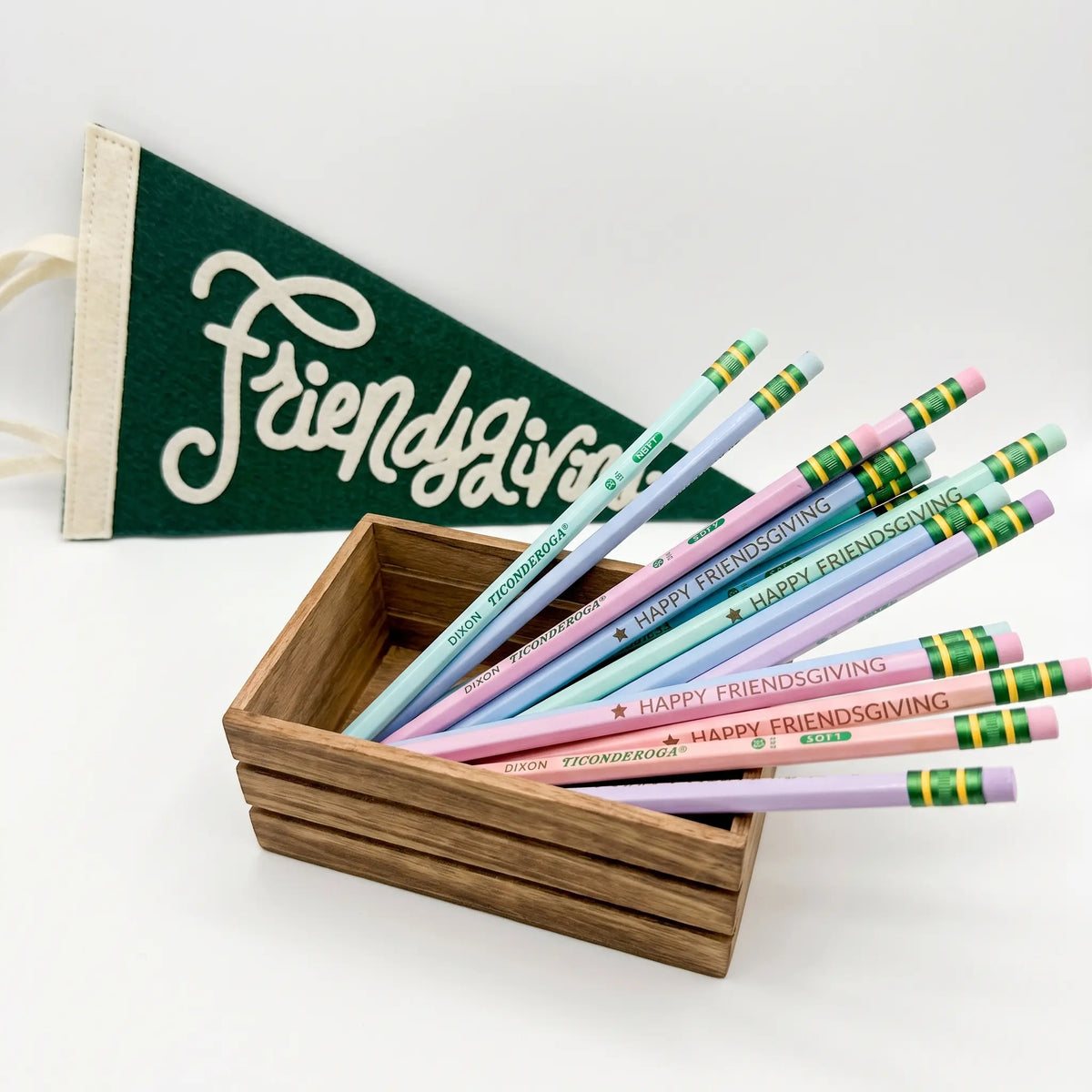 A set of pastel-colored pencils with 'Happy Friendsgiving' printed on them, displayed in a small wooden box next to a green felt pennant flag with white script reading 'Friends Giving'.