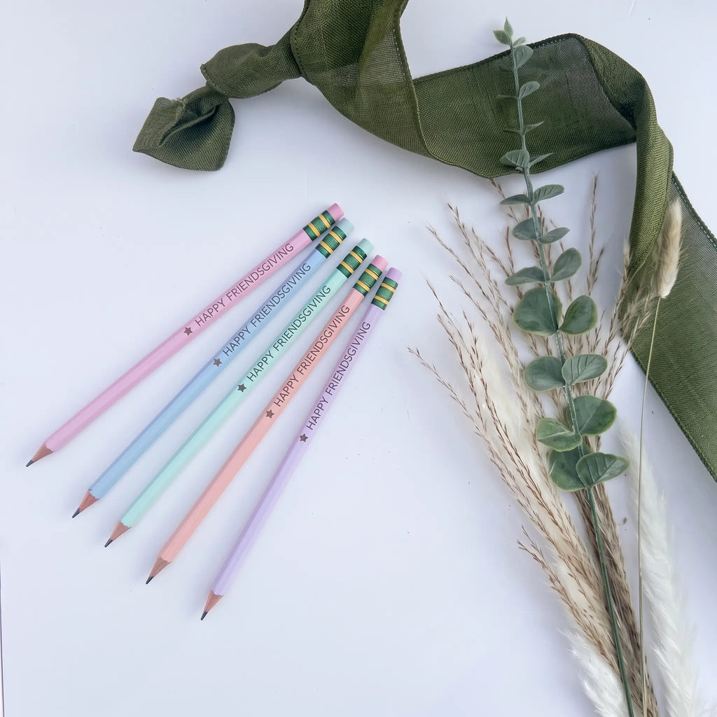 Five pastel-colored pencils with 'Happy Friendsgiving' messages and plaid erasers, arranged on a white surface with a green ribbon and dried grass stems.