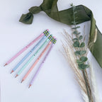 Five pastel-colored pencils with 'Happy Friendsgiving' messages and plaid erasers, arranged on a white surface with a green ribbon and dried grass stems.