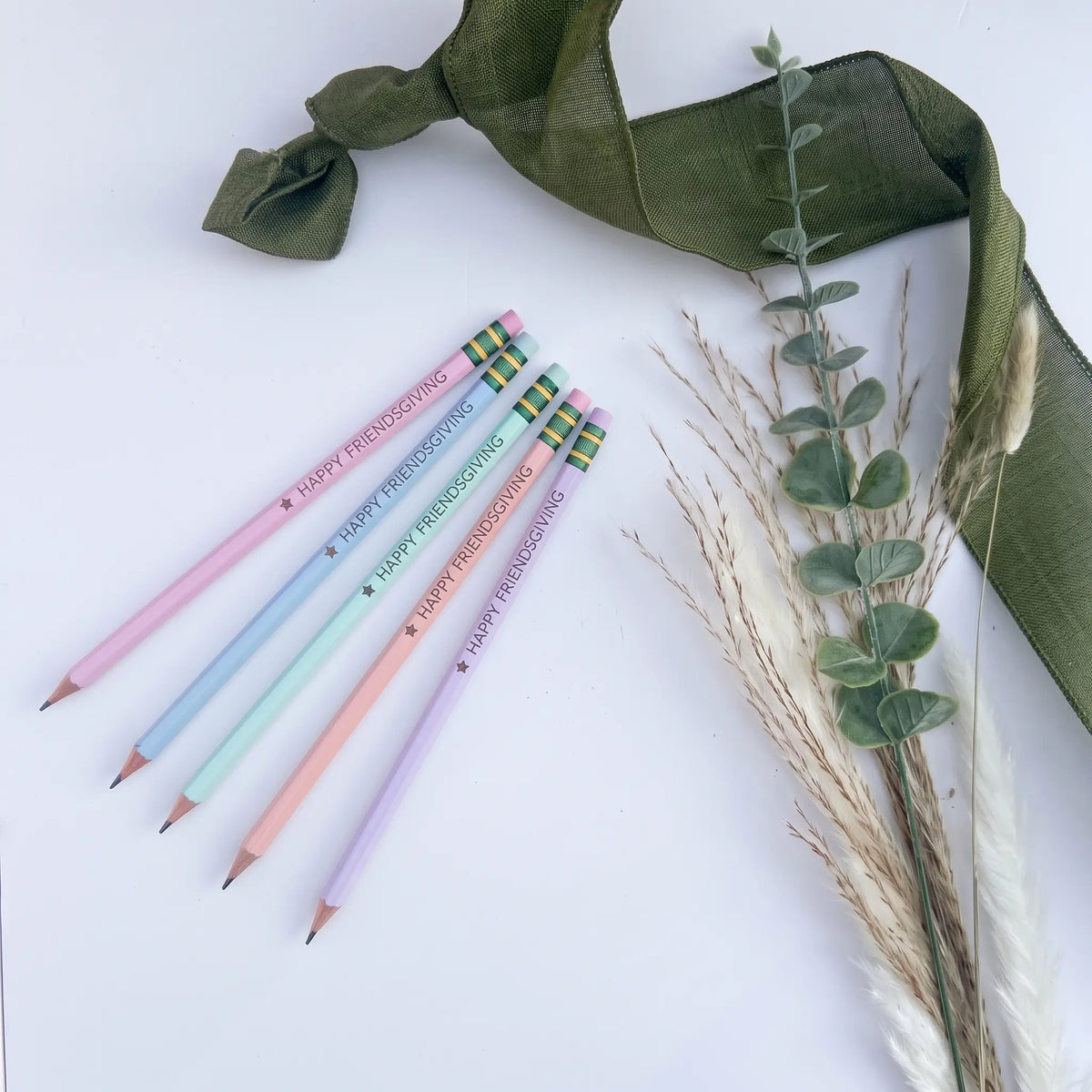 Five pastel-colored pencils with 'Happy Friendsgiving' messages and plaid erasers, arranged on a white surface with a green ribbon and dried grass stems.