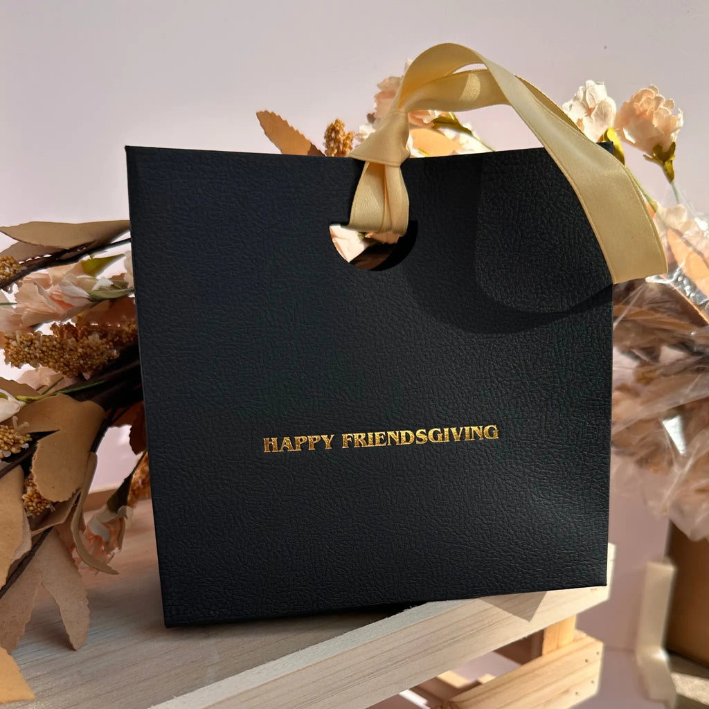 A black textured gift bag with gold foil lettering that reads 'HAPPY FRIENDSGIVING' and a gold ribbon handle, displayed on a wooden stand with dried floral decorations.