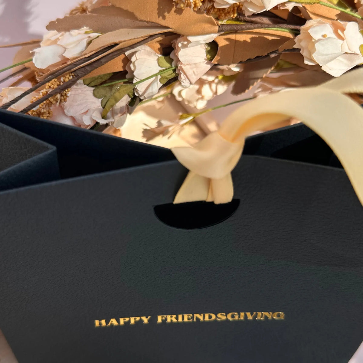 A close-up of a black gift bag with gold foil text reading 'HAPPY FRIENDSGIVING' and a cream ribbon tied around the handle, with dried flowers and brown leaves visible above.