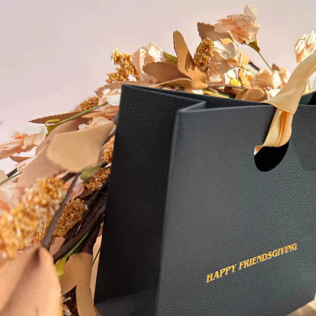 A close-up of a black textured paper gift bag with gold foil text reading 'HAPPY FRIENDSGIVING' and a beige ribbon handle, styled with dried beige flowers against a soft light background.
