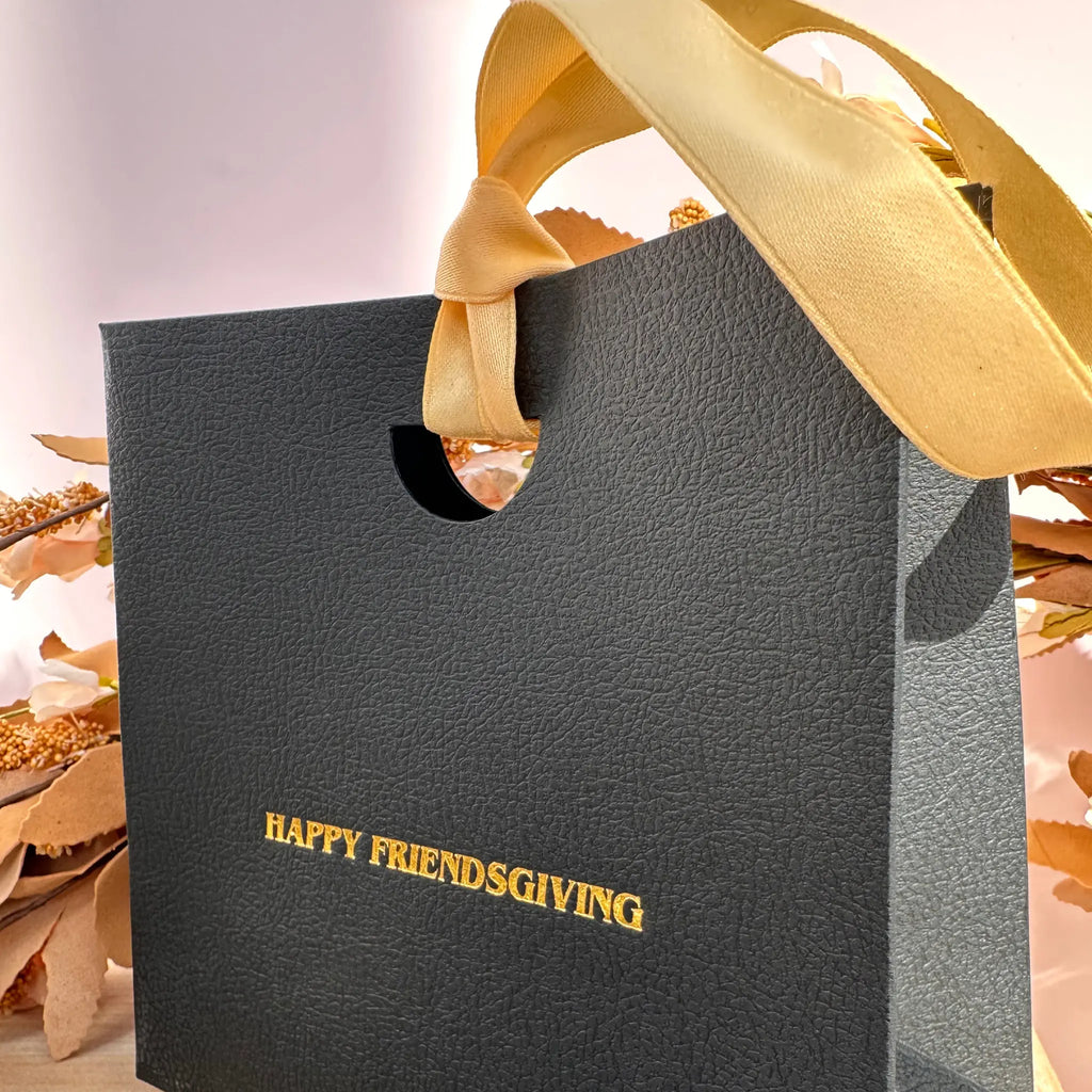 A textured black gift bag with a gold ribbon handle and gold foil text reading 'HAPPY FRIENDSGIVING' on the front, styled with dried autumn leaves against a soft neutral background.