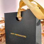 A textured black gift bag with a gold ribbon handle and gold foil text reading 'HAPPY FRIENDSGIVING' on the front, styled with dried autumn leaves against a soft neutral background.