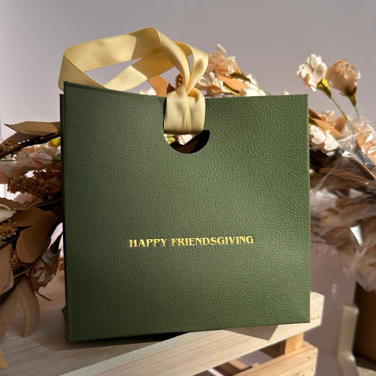 A textured olive green gift bag with a cream ribbon handle and gold foil lettering that reads 'HAPPY FRIENDSGIVING', displayed on a wooden surface with dried flowers in the background.