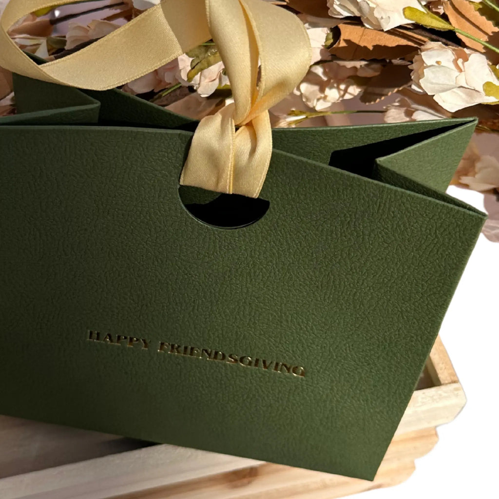 A dark green textured paper gift bag with a cream satin ribbon handle and embossed gold text reading 'HAPPY FRIENDSGIVING', shown on a wooden tray with dried flowers in the background.