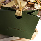 A dark green textured paper gift bag with a cream satin ribbon handle and embossed gold text reading 'HAPPY FRIENDSGIVING', shown on a wooden tray with dried flowers in the background.