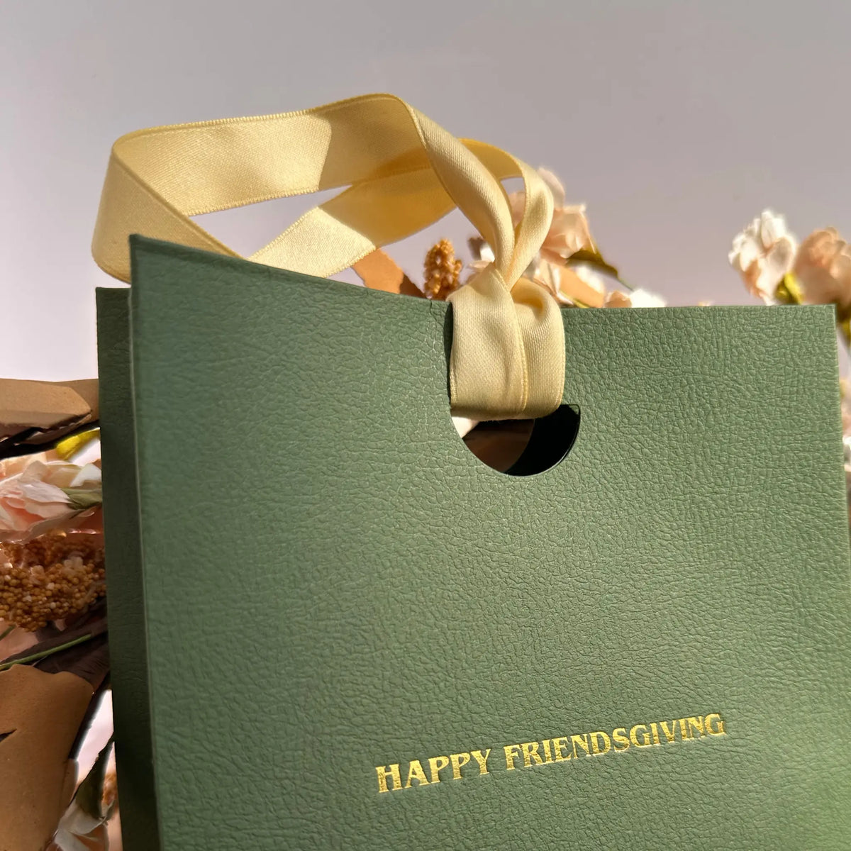 A textured sage green gift bag with a cream satin ribbon handle and gold foil text reading 'HAPPY FRIENDSGIVING' on the front, shown with dried flowers in the background.