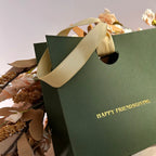 A dark green textured gift bag with gold foil text reading 'HAPPY FRIENDSGIVING' and cream satin handles, styled with dried flowers against a soft neutral background.