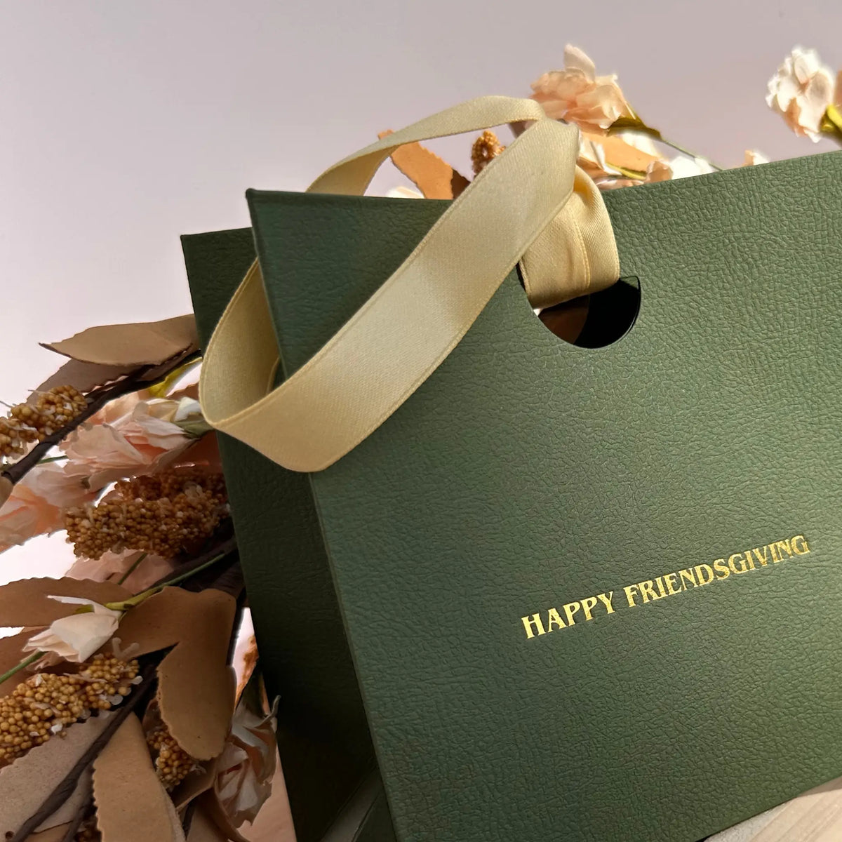 A dark green textured gift bag with gold foil text reading 'HAPPY FRIENDSGIVING' and cream satin handles, styled with dried flowers against a soft neutral background.