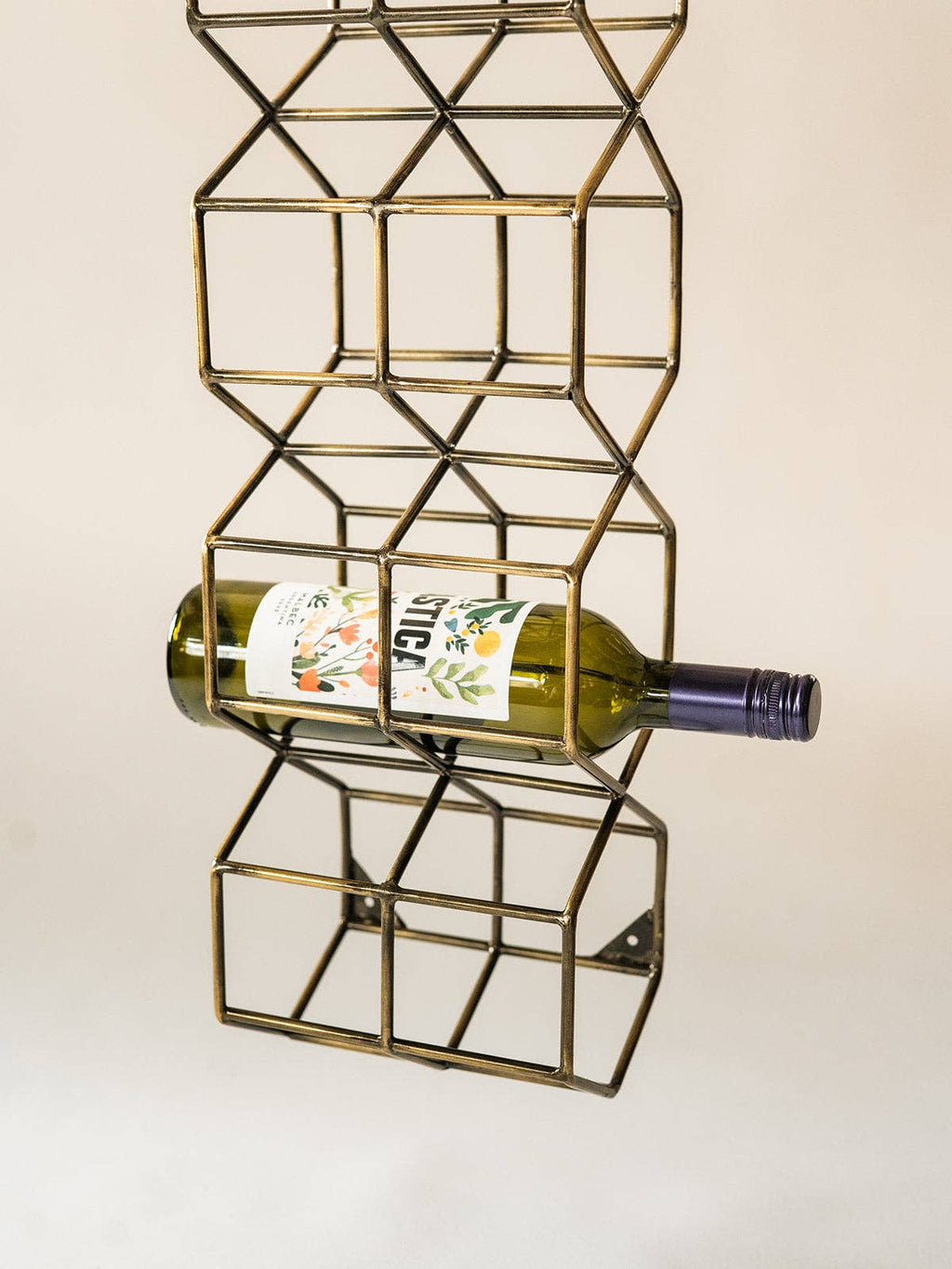 A vertical brass wire wine bottle rack with a geometric cube design, holding a green wine bottle horizontally against a plain off-white background.