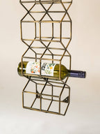 A vertical brass wire wine bottle rack with a geometric cube design, holding a green wine bottle horizontally against a plain off-white background.
