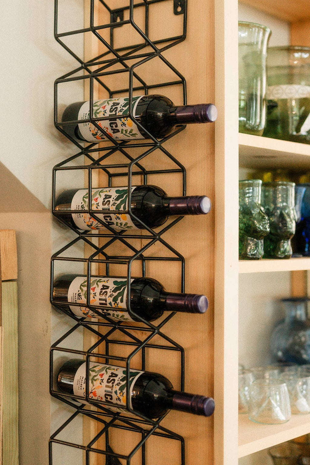 A black metal wire wine rack mounted vertically on a light wood cabinet door, holding four wine bottles horizontally. To the right are light wood shelves with glassware.