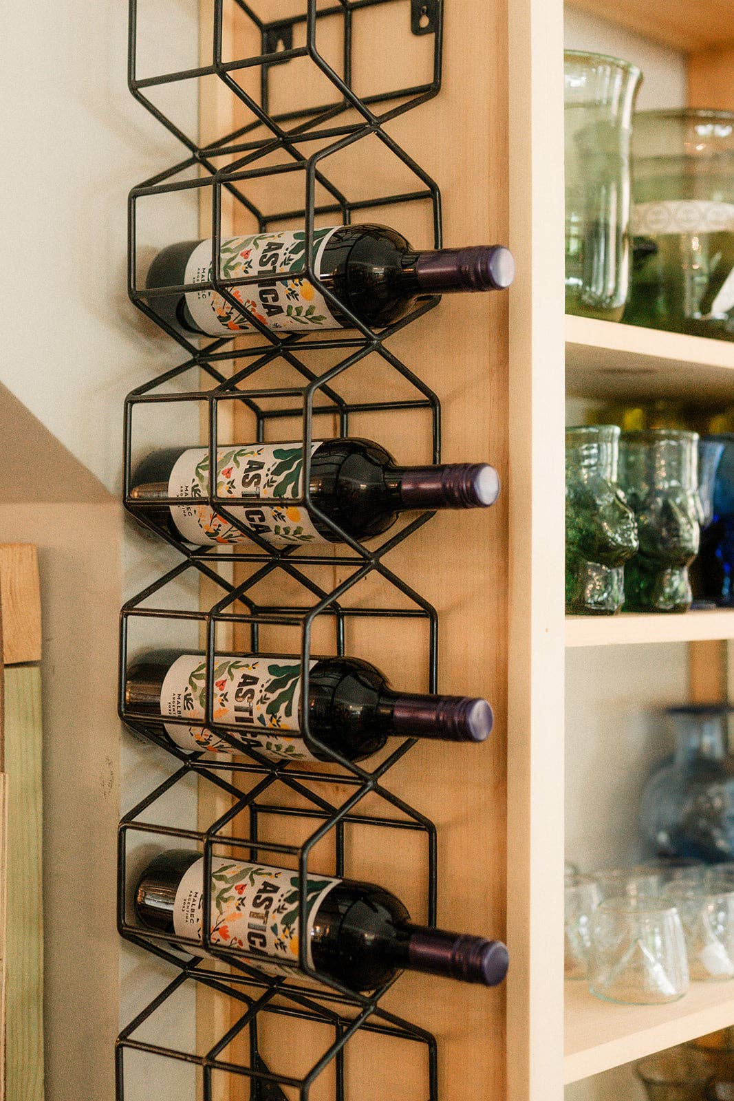 A black metal wire wine rack mounted vertically on a light wood cabinet door, holding four wine bottles horizontally. To the right are light wood shelves with glassware.
