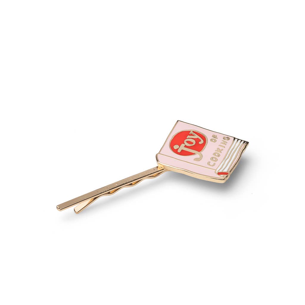 A gold-tone hair clip shaped like a book with a light pink cover, featuring a red oval logo with the word 'joy' and the phrase 'the joy of cooking' in gold lettering.
