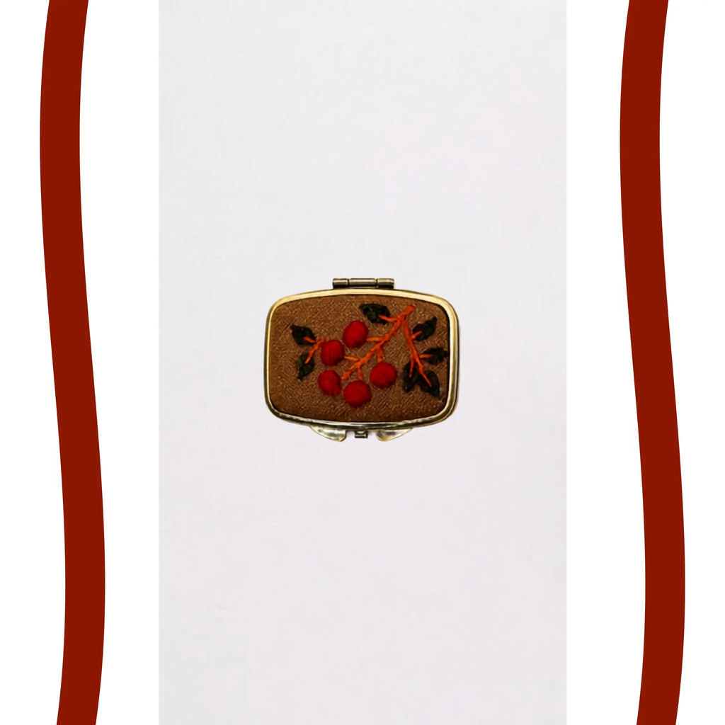 A small rectangular gold-toned case or box with a brown textured cover, decorated with an embroidered or painted red berry motif and dark green leaves, centered on a white background with decorative red side bars.