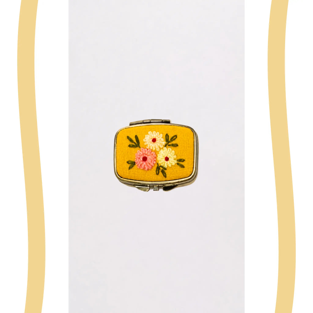 A small vintage-style compact case with a yellow floral enamel face, featuring embroidered flowers in pink, yellow, and white with green leaves, set against a white background with decorative gold wavy borders on the sides.