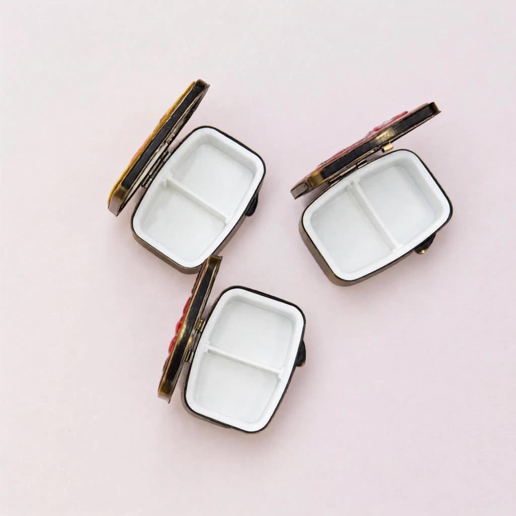 Three small rectangular compact mirrors with white divided interiors and dark metal edges, arranged on a light pink background.