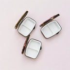 Three small rectangular compact mirrors with white divided interiors and dark metal edges, arranged on a light pink background.