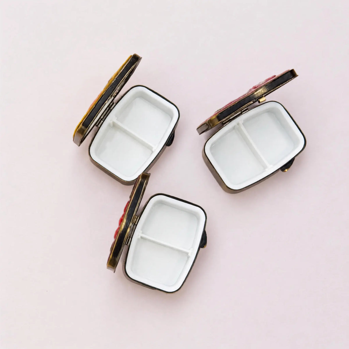 Three small rectangular compact mirrors with white divided interiors and dark metal edges, arranged on a light pink background.