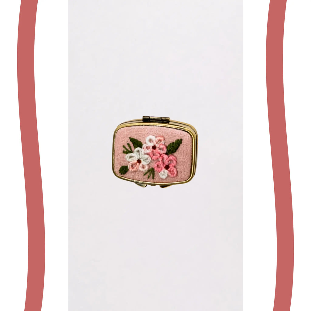 A small rectangular pink clutch or compact case with gold-tone metal edges and an embroidered floral design featuring pink, white, and green flowers on a plain white background.