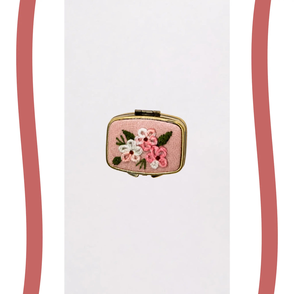 A small rectangular pink clutch or compact case with gold-tone metal edges and an embroidered floral design featuring pink, white, and green flowers on a plain white background.