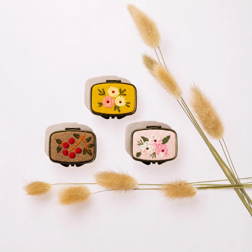 Three small embroidered patchwork cases with a cream-colored fabric exterior and colorful floral and berry embroidery, arranged on a white background with dried grass stems.